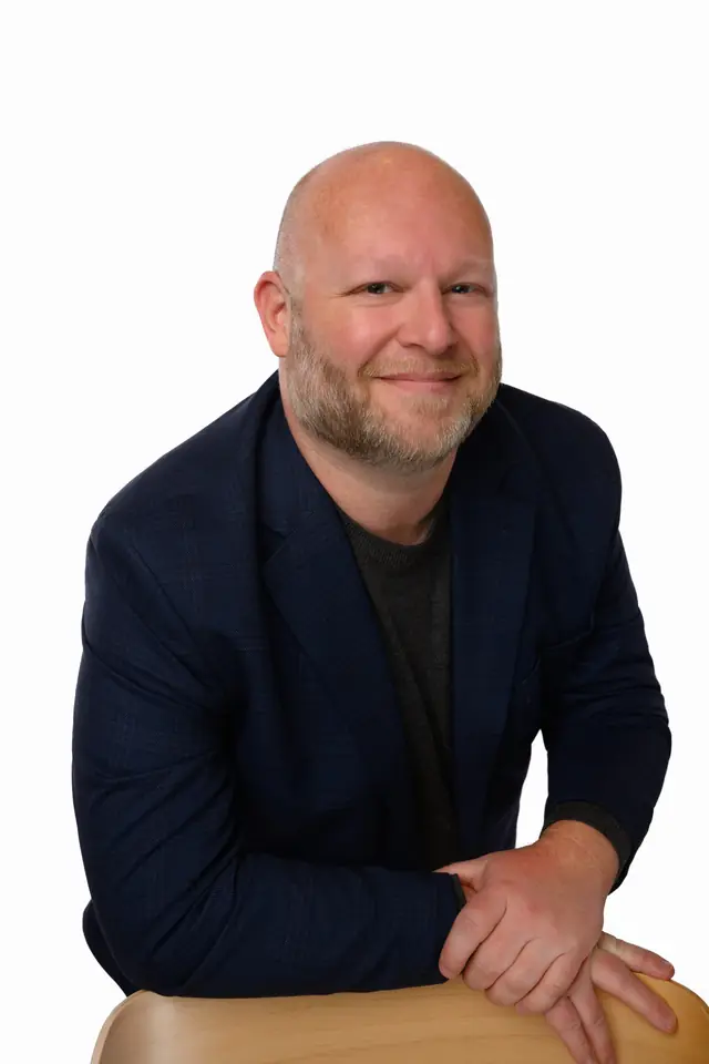 Portrait of Kristopher Jenkins in a navy blazer, smiling, leaning on a chair back